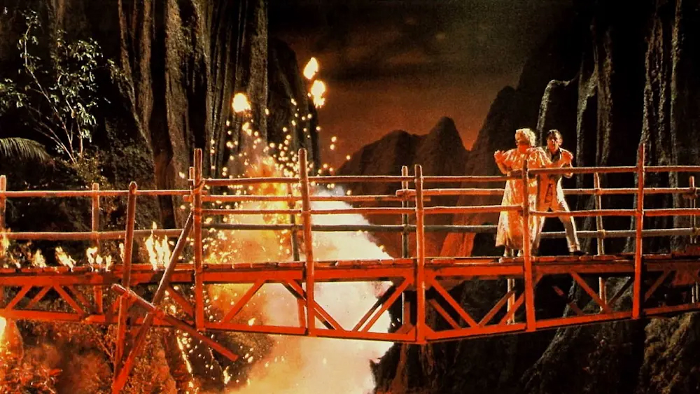 The bridge scene in When Time Ran Out... showing a rickety bridge being crossed by a scared looking man and woman, while a pillar of smoke and sparks bursts up from the gorge below. All is red-lit from the stream of lava flowing through the gorge unseen beneath
